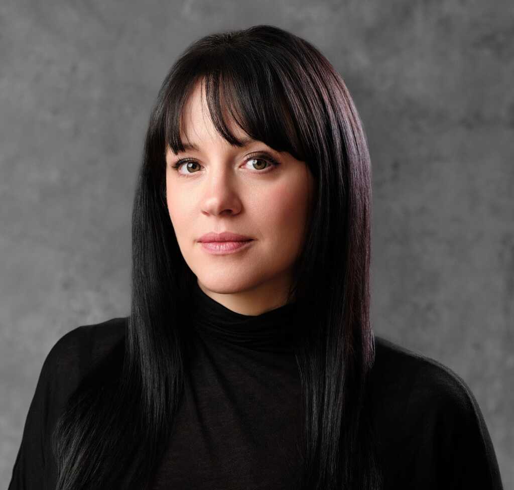 A person with long black hair and bangs is wearing a black top, looking directly at the camera against a gray background.