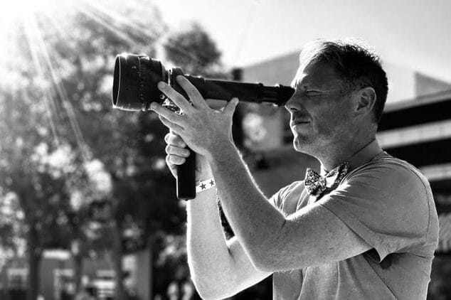 A man in a short-sleeve shirt holds and looks through a vintage camera outdoors in bright sunlight, capturing a moment of Silver Screen filmmaking; blurred trees and buildings linger in the black-and-white background.