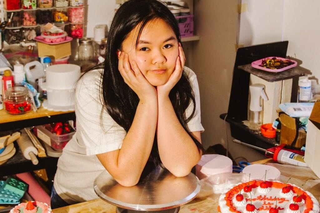 A young woman with long dark hair rests her chin on her hands and smiles while standing behind a table with several decorative, strawberry-topped cakes in a colorful, cluttered kitchen or workspace.