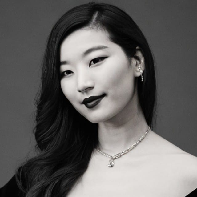 A young woman with long, wavy hair poses against a plain background, embodying the poise of someone pursuing a creative career. She wears dark lipstick, a necklace, and an ear cuff. Her calm, confident expression is captured in black and white.