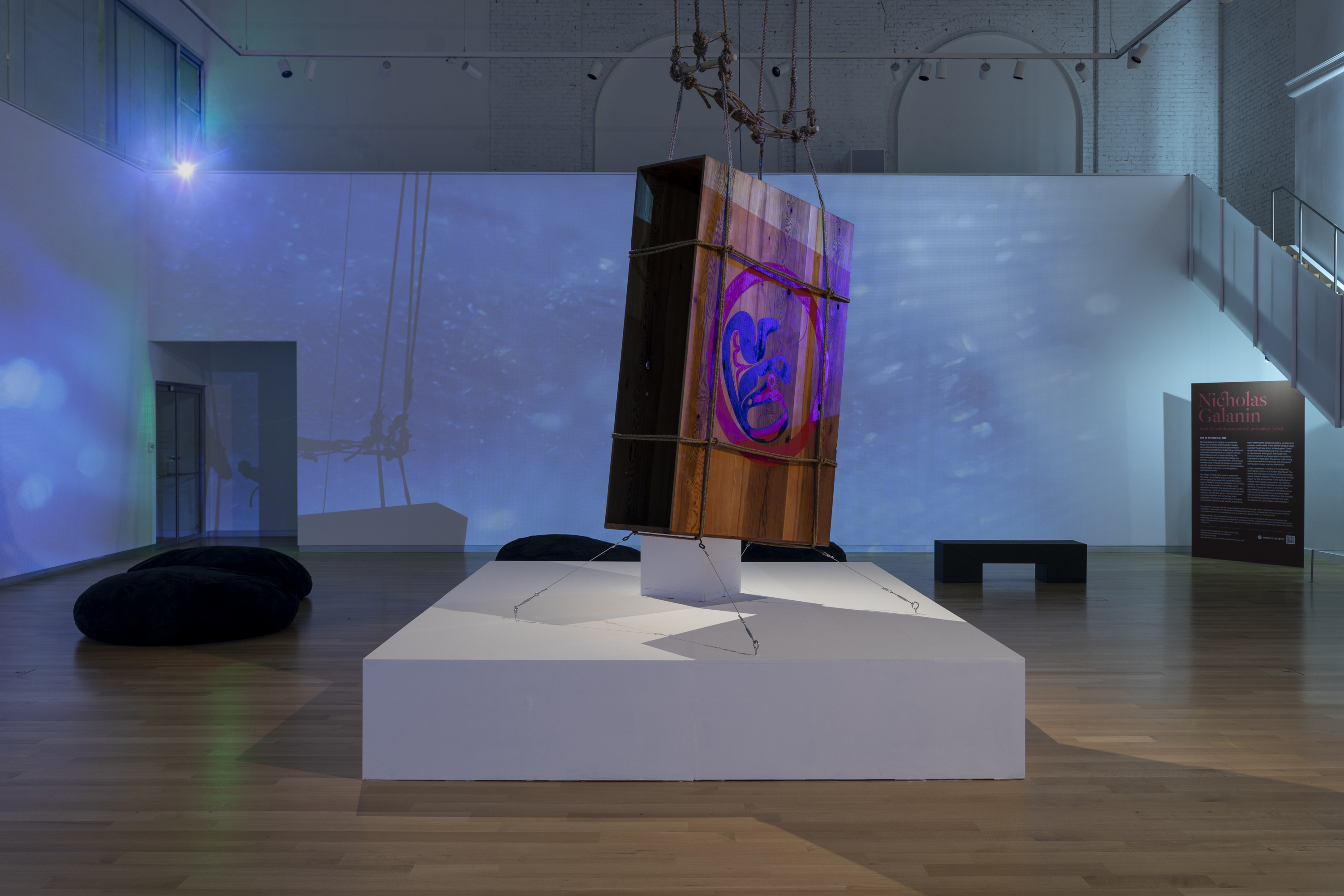 A large, weathered wooden box with a painted purple G hangs tilted on a white platform in a modern gallery, with blue light projections and a red information panel in the background.