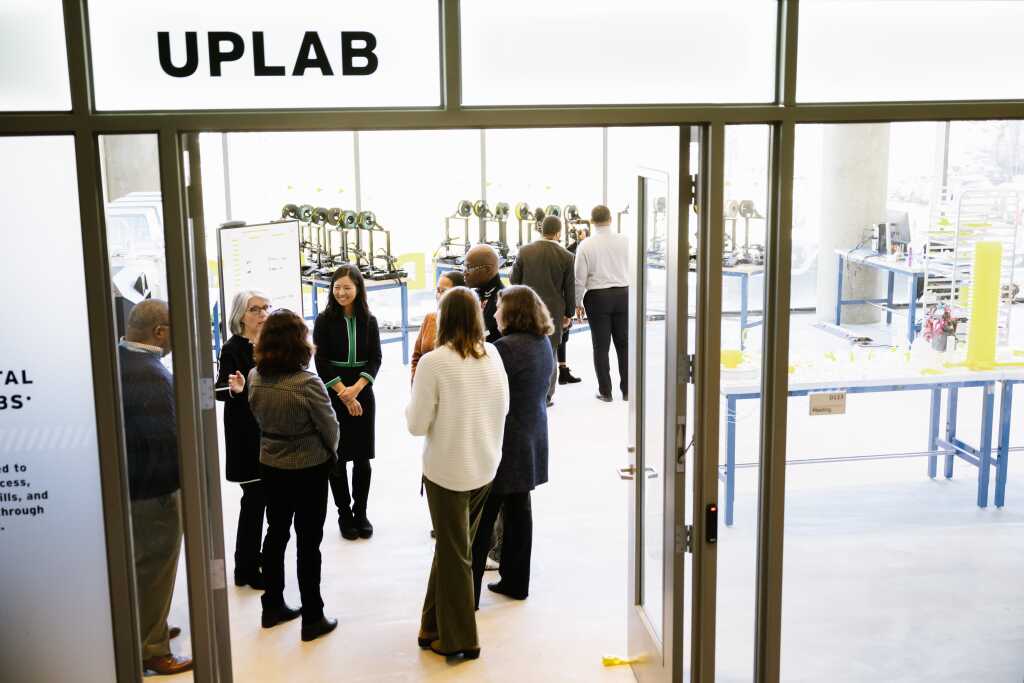 A group of people stands and talks inside a modern lab space labeled “UPLAB,” with studios, equipment, and tables visible in the background through large windows, highlighting the collaborative resources available.