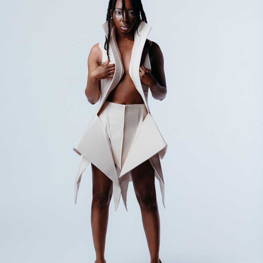 A woman stands confidently in a unique, beige geometric outfit reminiscent of MassArt style, with an open vest and angular skirt, paired with high-heeled sandals, against a plain white background.
