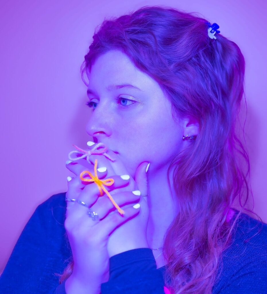 A young woman with long hair gazes thoughtfully to the side, holding her hands near her face. She has white nail polish, rings, and wears bows on her fingers—an evocative illustration lit with purple and blue hues.