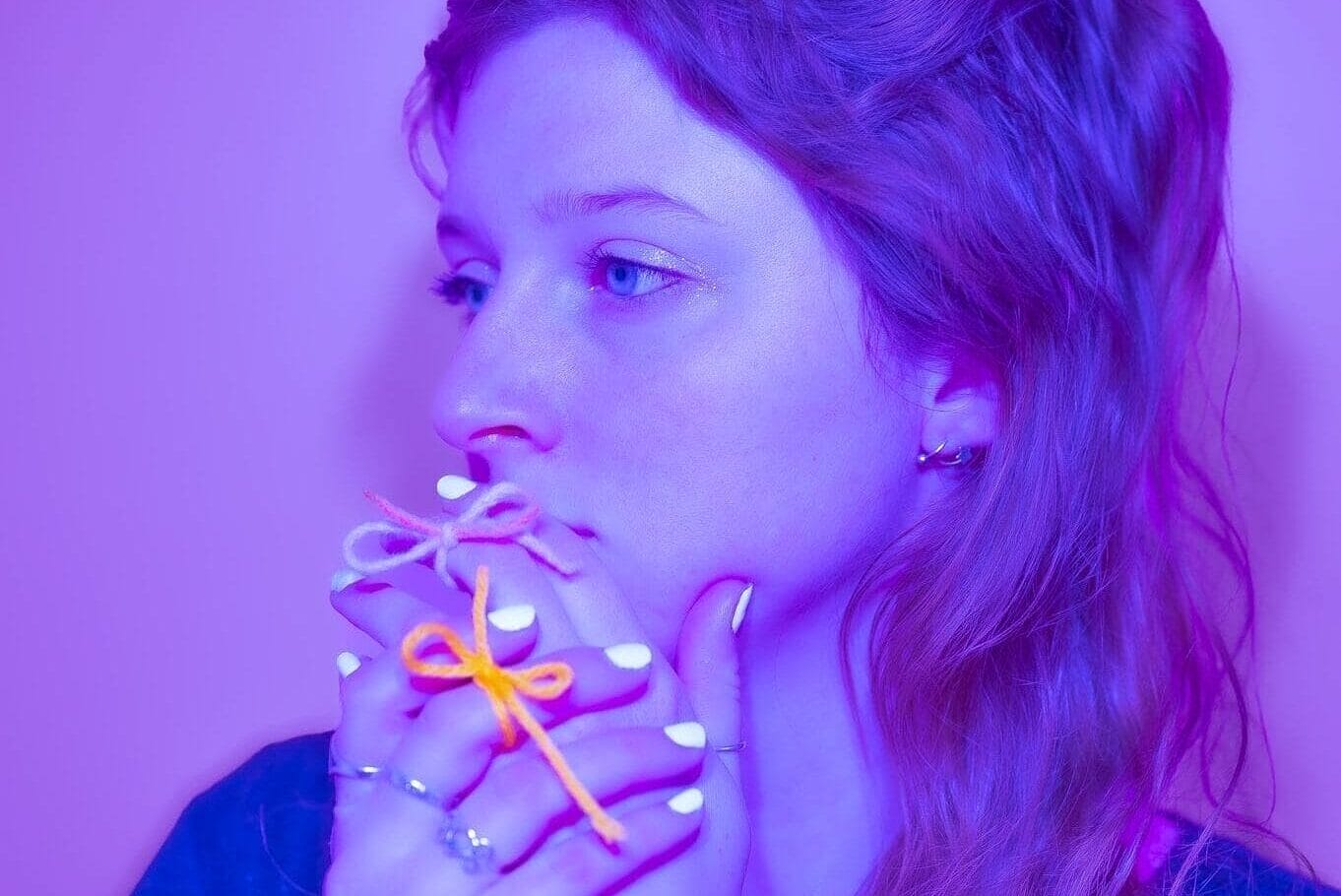 A young woman with long hair gazes thoughtfully to the side, holding her hands near her face. She has white nail polish, rings, and wears bows on her fingers—an evocative illustration lit with purple and blue hues.