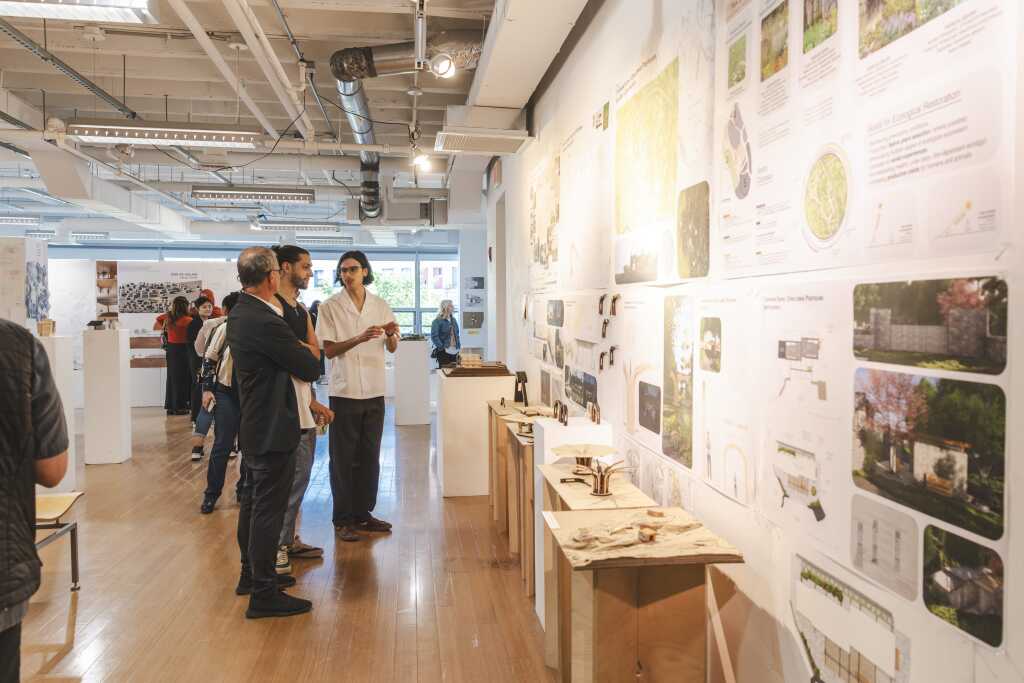 People view architectural models and design boards displayed on a brightly lit gallery wall at MassArt. Others explore the exhibition space in the background, with shelves, spotlights, and a modern, open-ceiling interior.