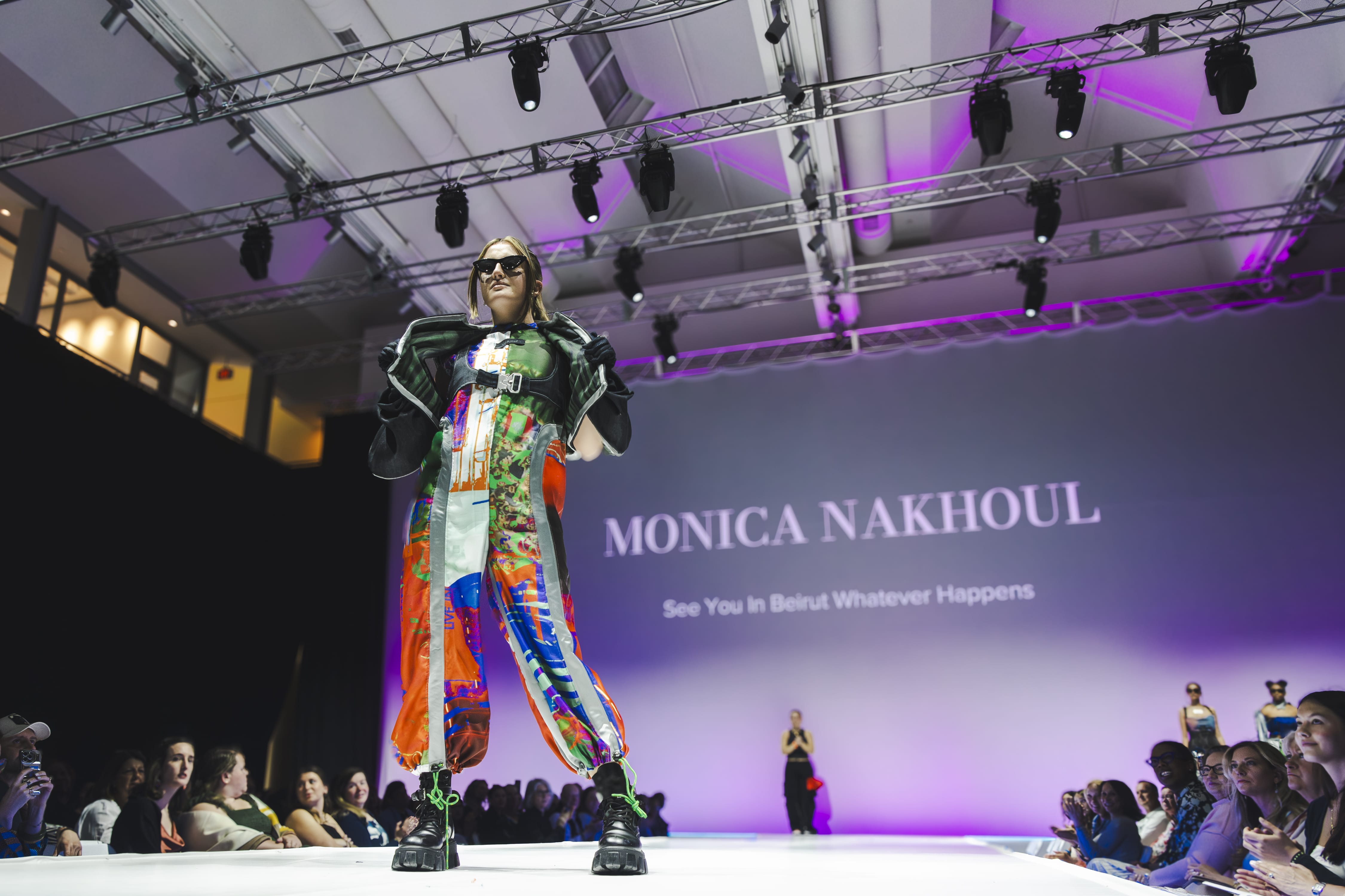 A model wearing colorful, abstract-patterned clothing and sunglasses walks down a brightly lit runway at the Fashion Show, with MONICA NAKHOUL and See You in Beirut Whatever Happens projected on the wall behind. Audience watches from both sides.
