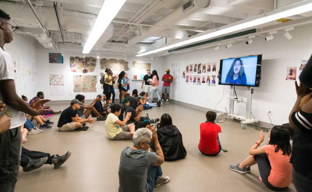 A diverse group of people sit on the floor and stand in a brightly lit art gallery, prioritizing the speaker on a large TV screen as colorful artwork decorates the white walls around them.