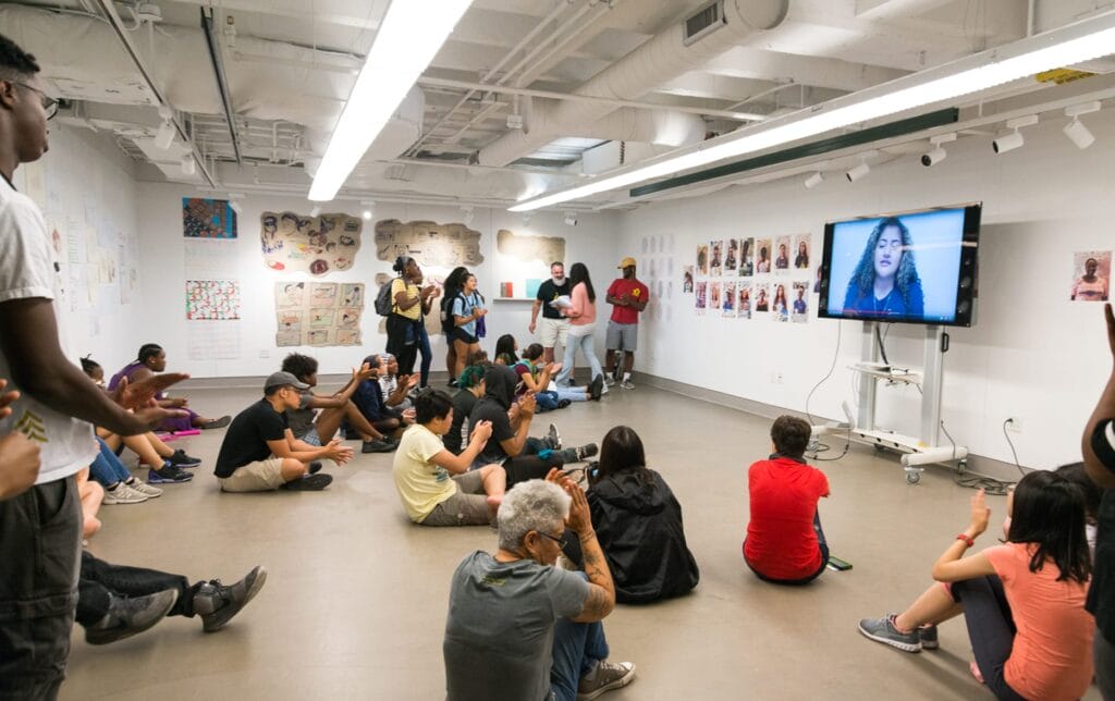 A diverse group of people sit on the floor and stand in a brightly lit art gallery, prioritizing the speaker on a large TV screen as colorful artwork decorates the white walls around them.