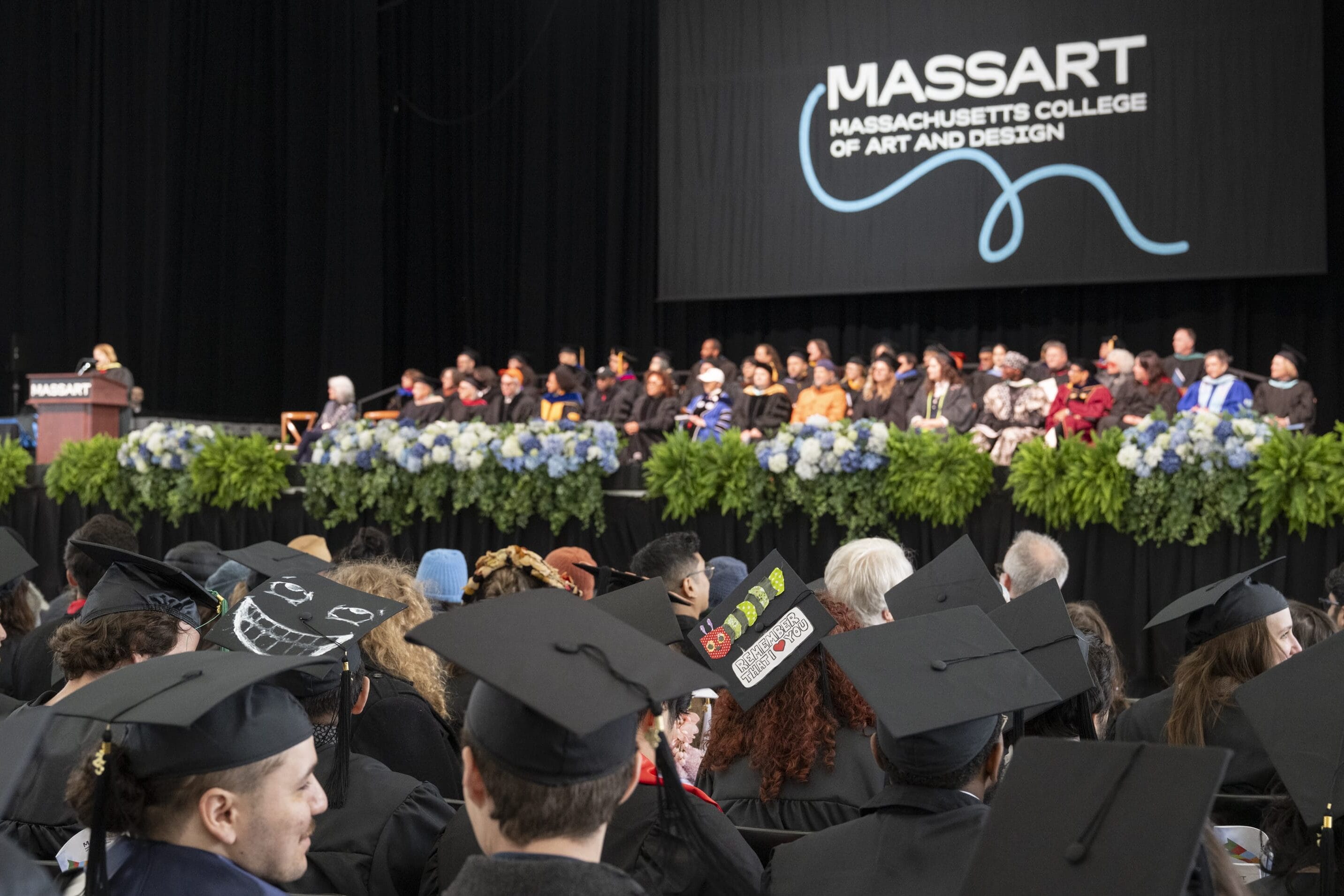 Graduates in caps and gowns sit facing a stage decorated with flowers, where faculty and speakers are seated under a large sign reading Massachusetts College of Art and Design during a commencement ceremony.