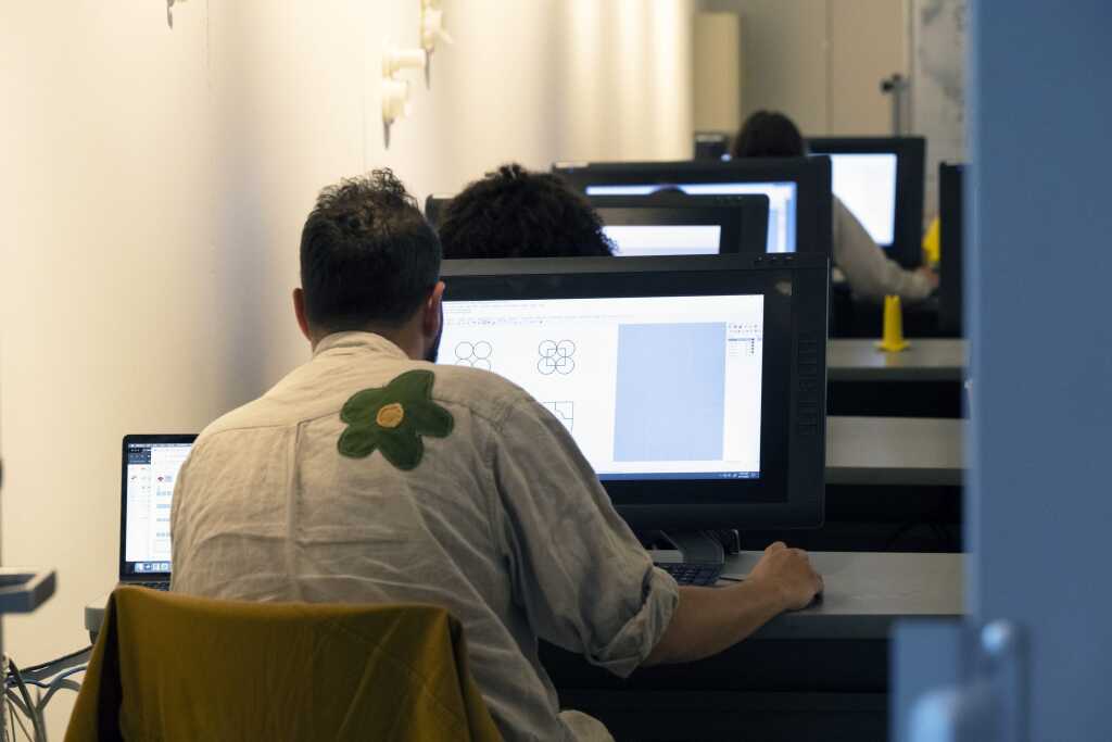 A person sits at a desk working on a computer in a classroom or office setting, with several others at computers in a row ahead of them. The persons shirt has a green flower design on the back, reflecting the creative energy often found in studios and shared resources.