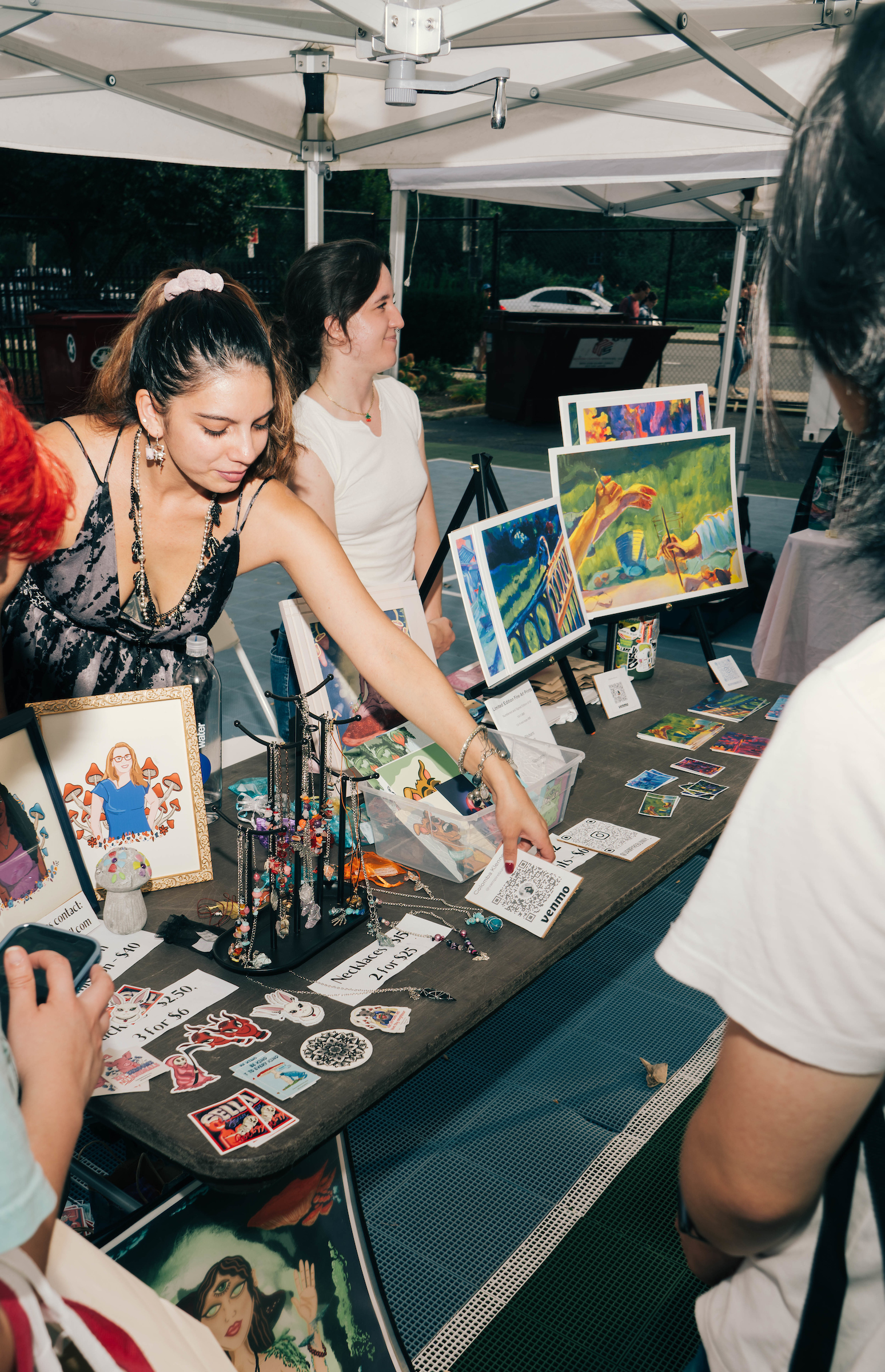 A woman at an outdoor art market booth gestures towards a QR code on a table displaying vibrant prints, stickers, and jewelry, proudly showcasing her MassArt creations as several people observe her work.