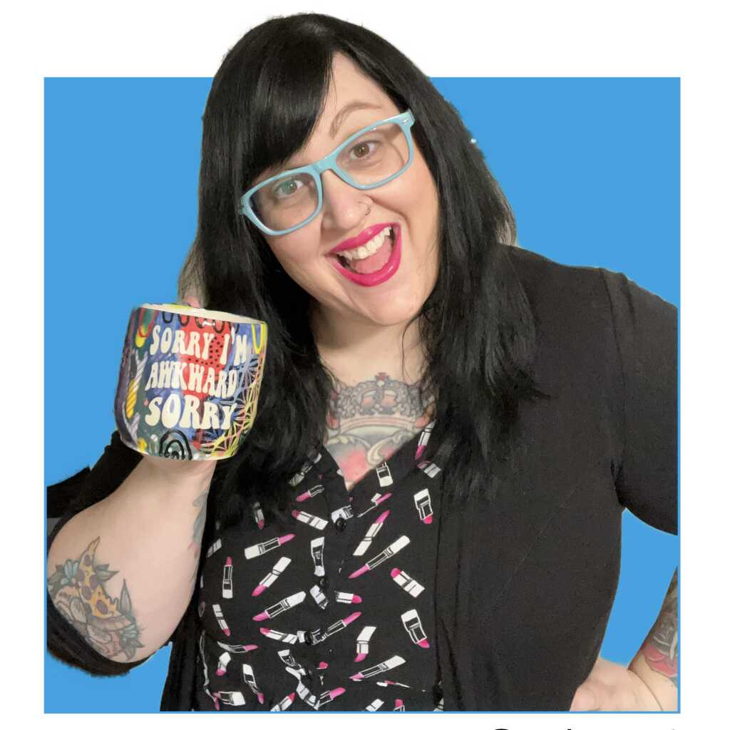 A smiling woman with long black hair and blue glasses holds up a colorful mug that says Sorry Im awkward. She has tattoos and wears a black cardigan over a dress with a lipstick print, standing against a blue background.