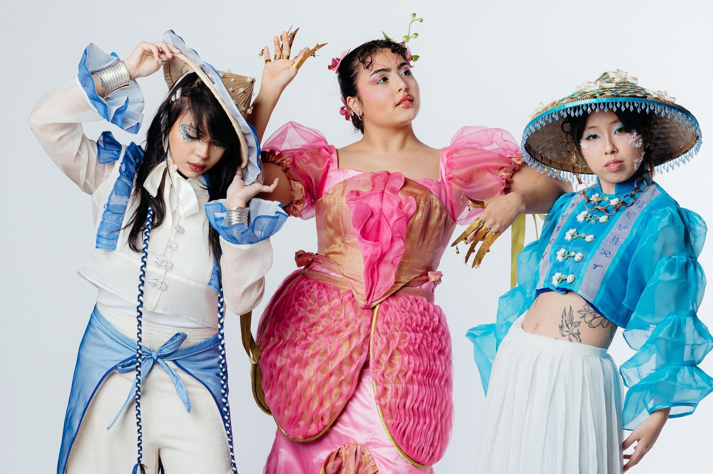 Three people pose as if ready for a Fashion Show: one in a white and blue ruffled ensemble, one in a pink flower-inspired gown with green accents, and one in a blue sheer top, white skirt, and wide-brimmed straw hat.