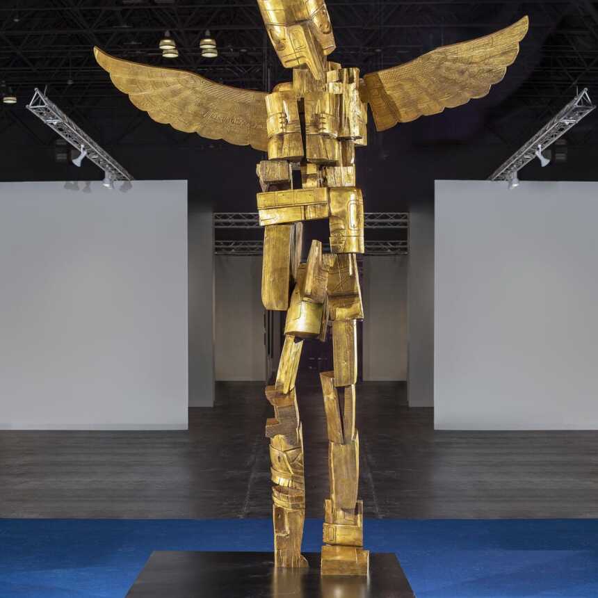 A tall gold sculpture of a humanoid figure with wings stands on a black platform in a spacious, modern indoor exhibition hall with blue flooring and overhead lights.