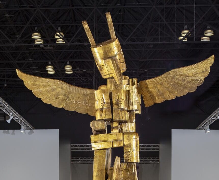 A tall gold sculpture of a humanoid figure with wings stands on a black platform in a spacious, modern indoor exhibition hall with blue flooring and overhead lights.