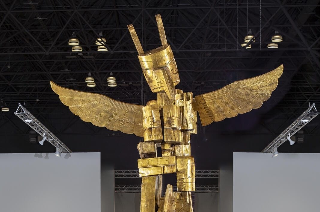 A tall gold sculpture of a humanoid figure with wings stands on a black platform in a spacious, modern indoor exhibition hall with blue flooring and overhead lights.