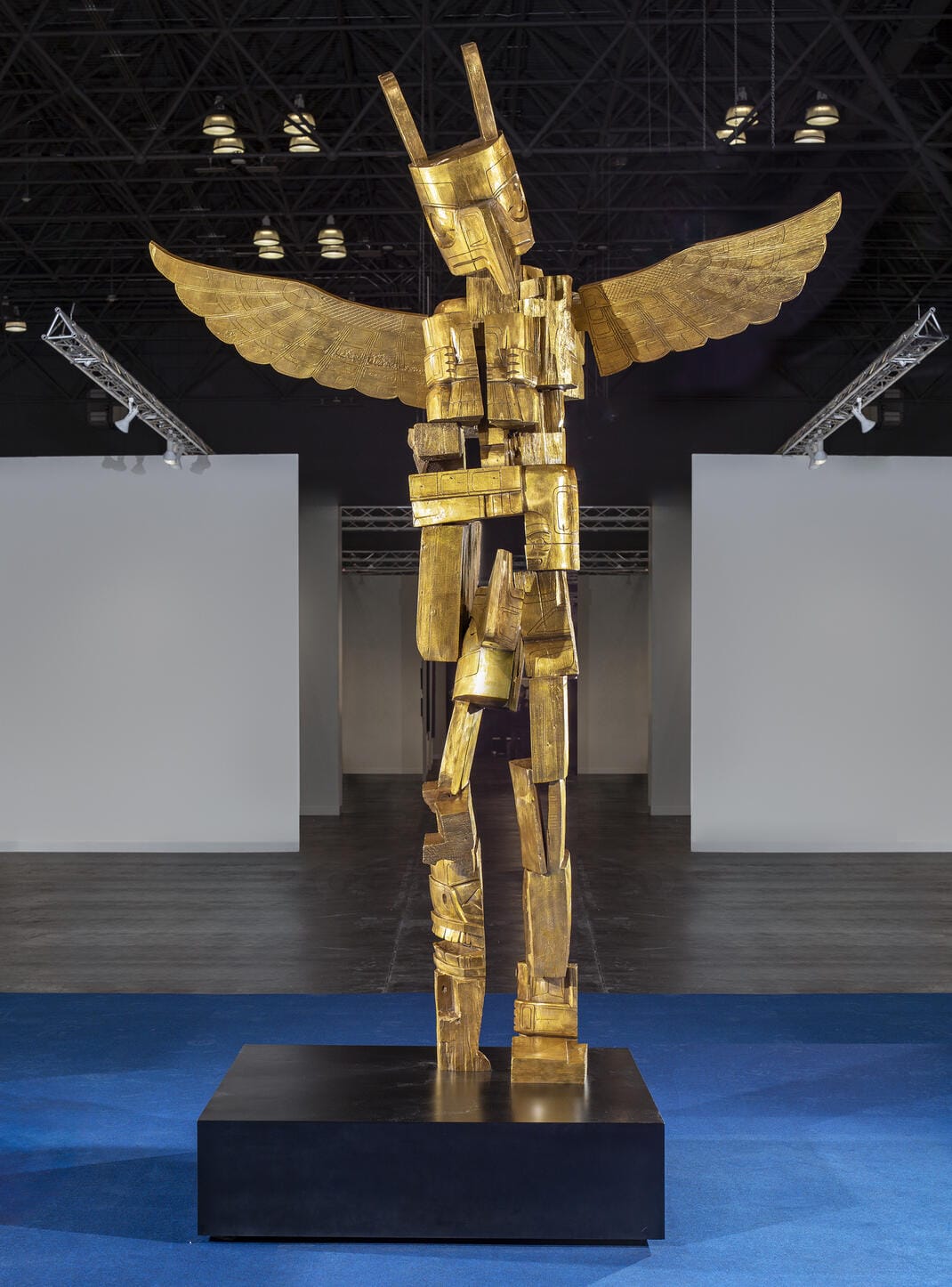 A tall gold sculpture of a humanoid figure with wings stands on a black platform in a spacious, modern indoor exhibition hall with blue flooring and overhead lights.