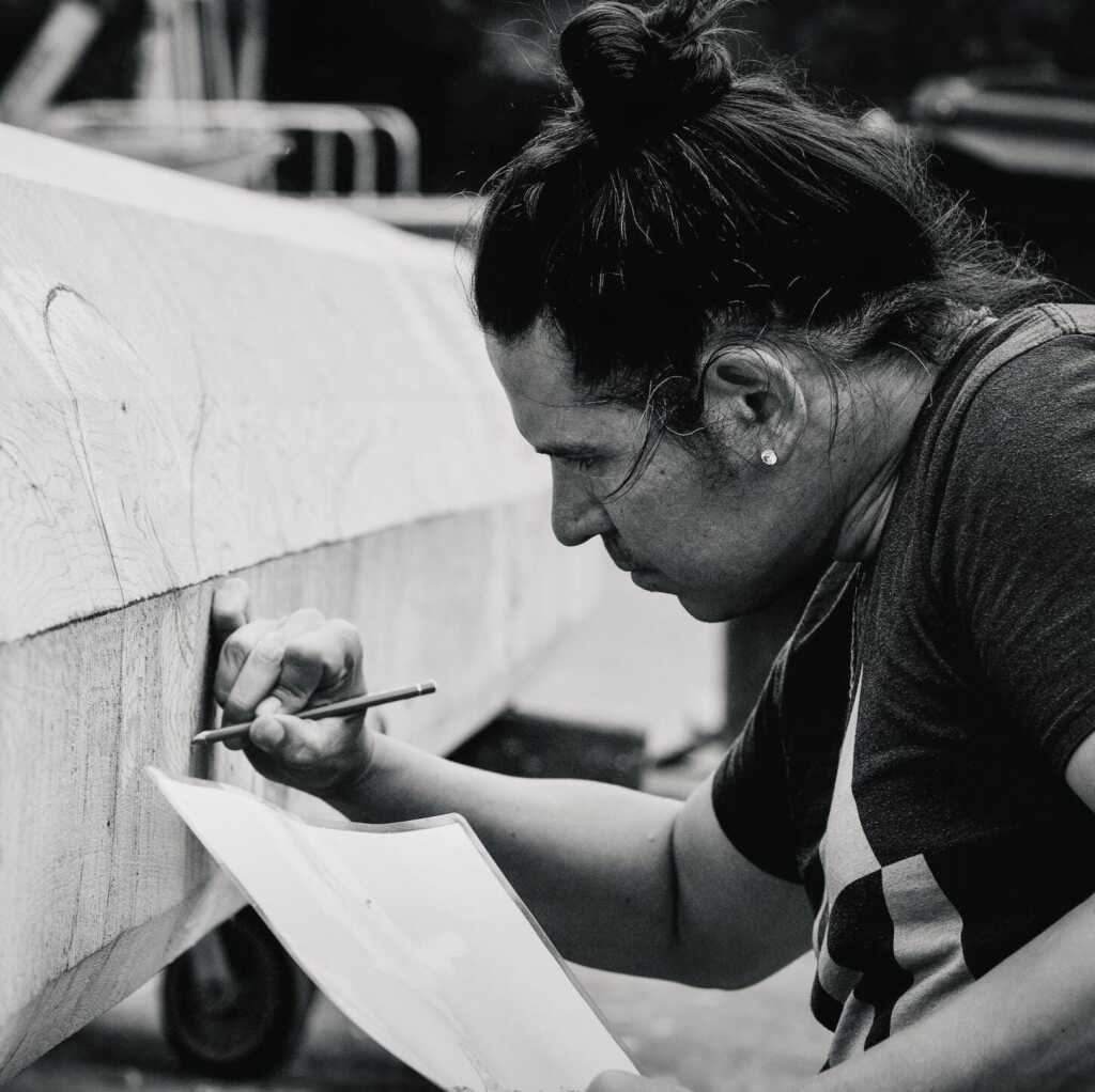 A person with long hair tied in a bun carefully draws or carves on a large wooden object at MassArt, holding paper in one hand and a pencil or tool in the other. The image is in black and white.