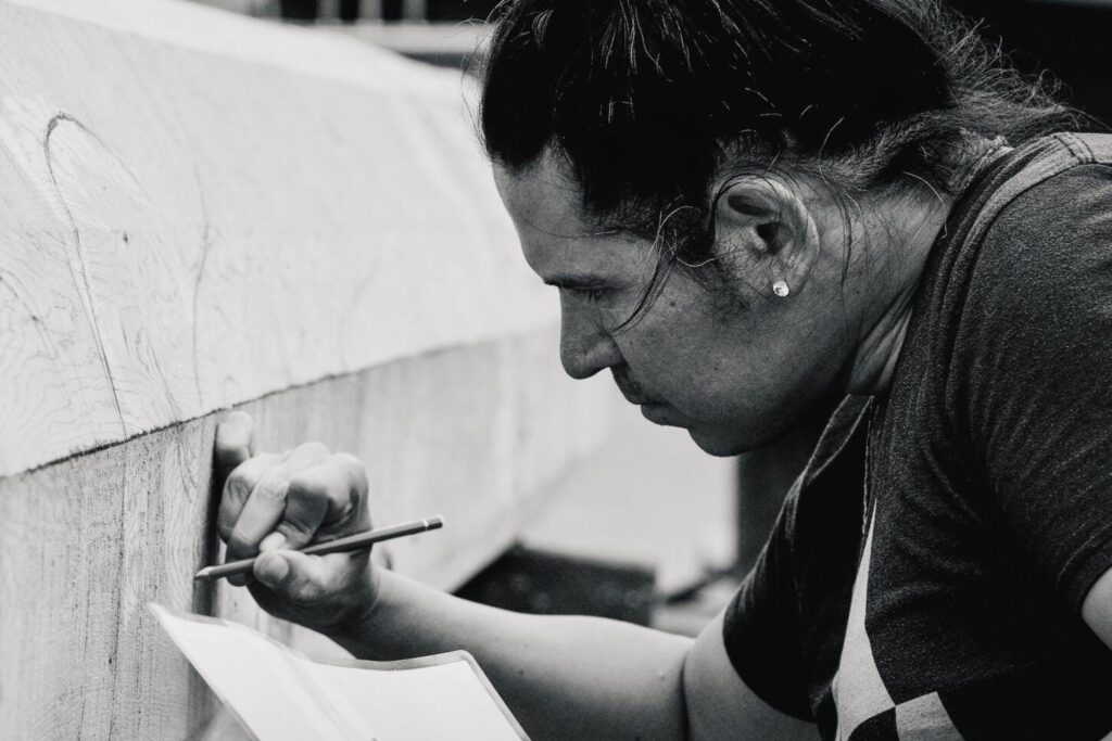A person with long hair tied in a bun carefully draws or carves on a large wooden object at MassArt, holding paper in one hand and a pencil or tool in the other. The image is in black and white.