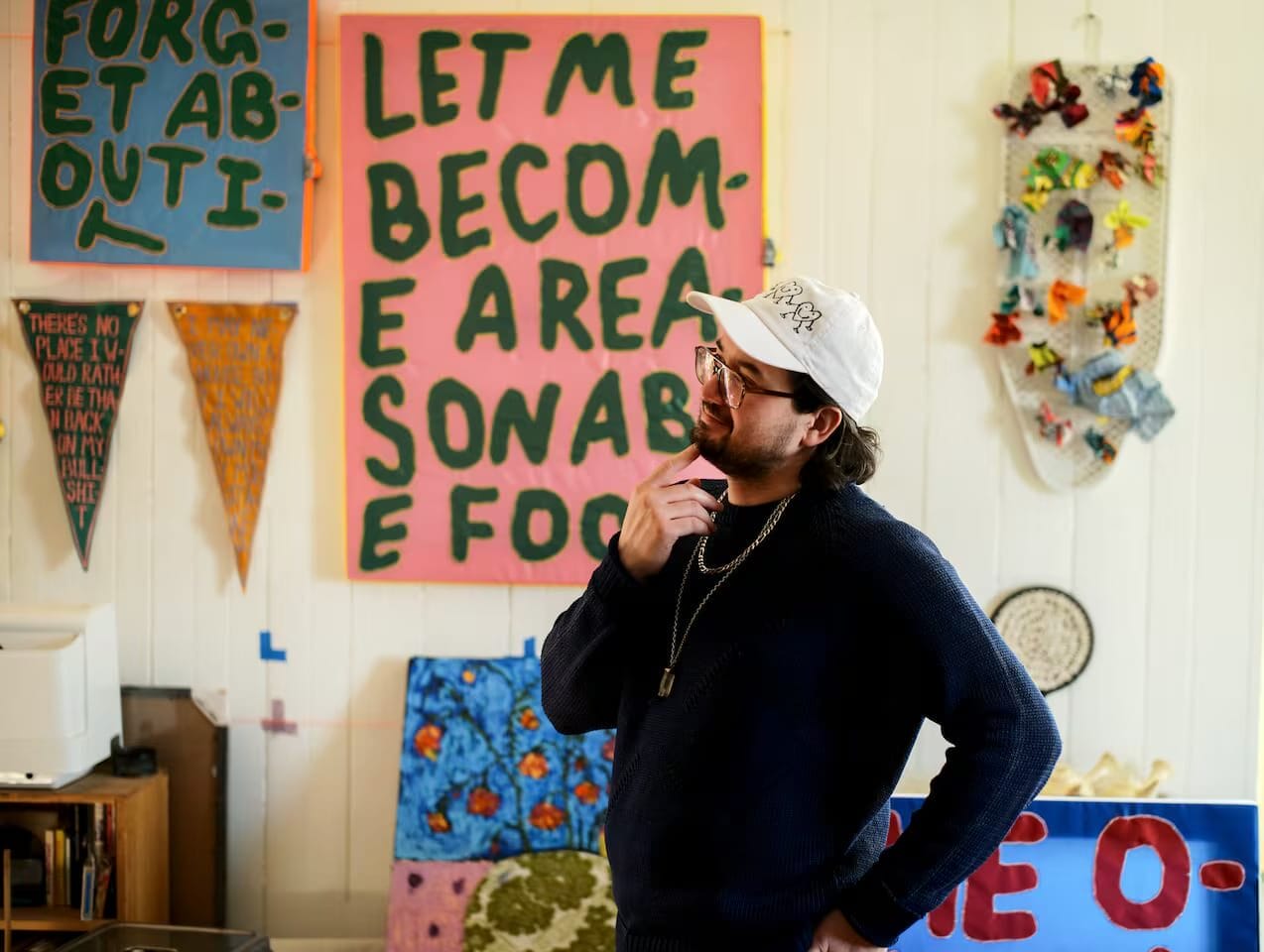 A person in a white cap and dark sweater stands thoughtfully in a colorful room adorned with sunny side hues and vibrant, hand-painted signs—artwork by artist Andy Li—including one that reads LET ME BECOME A REASONABLE FOOD.