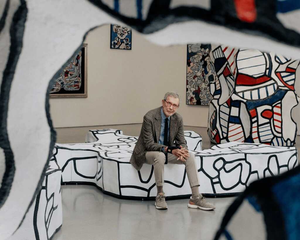 An art dealer sits in a blazer on abstract, white sculptural benches with black line art in an art gallery, surrounded by large, colorful artworks and sculptures—an inspiring setting for anyone interested in an art dealer career or art collecting.