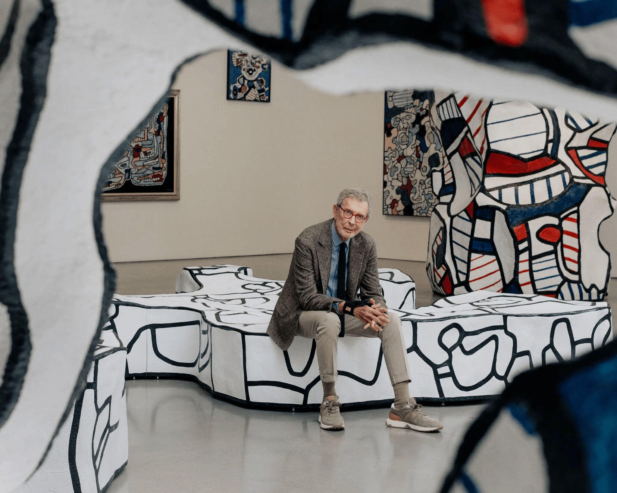 An art dealer sits in a blazer on abstract, white sculptural benches with black line art in an art gallery, surrounded by large, colorful artworks and sculptures鈥攁n inspiring setting for anyone interested in an art dealer career or art collecting.