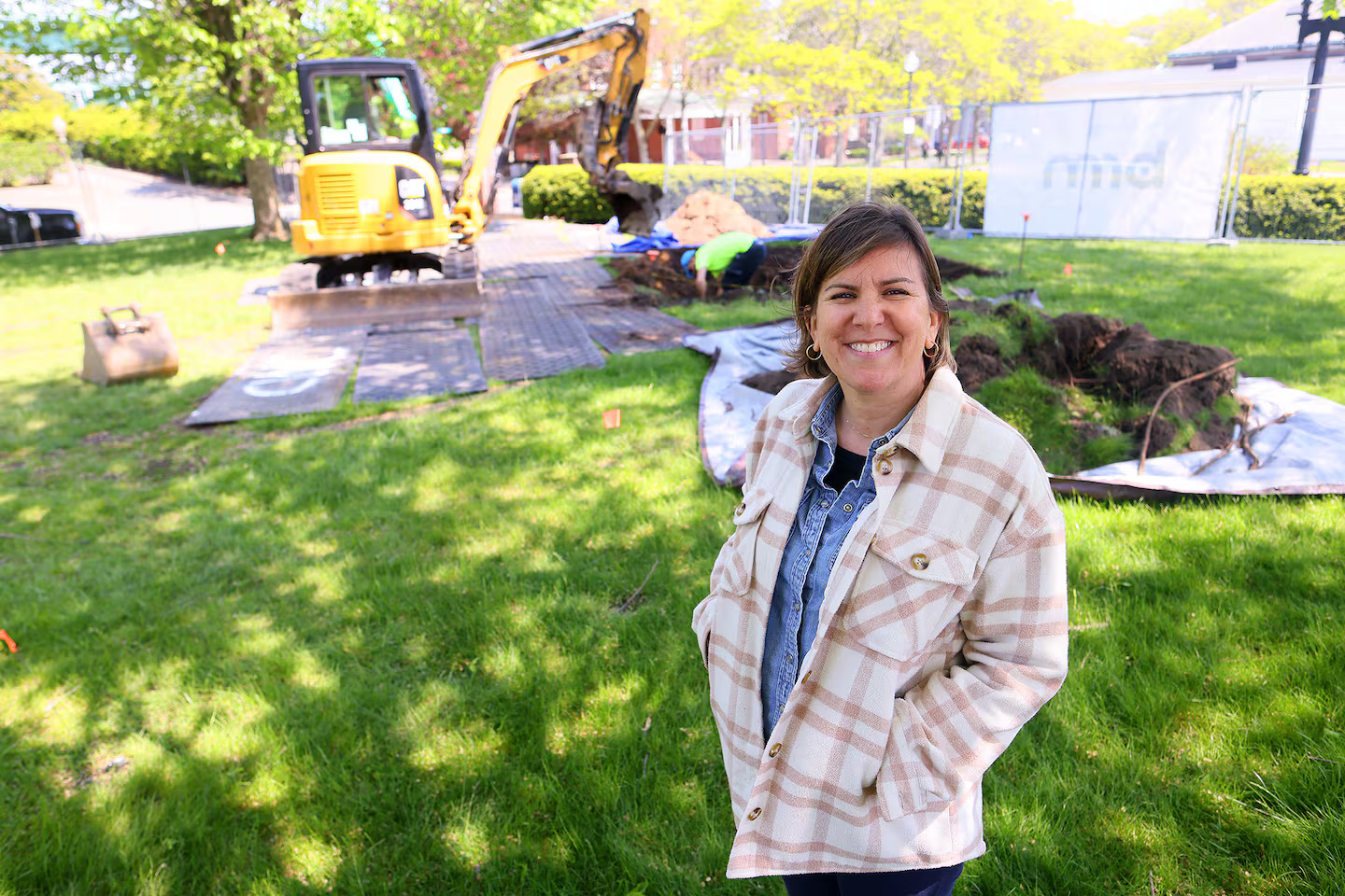 A woman in a plaid jacket smiles at the camera while standing on a grassy area; behind her, a small excavator and a Triennial artist working with soil are visible on a sunny day.