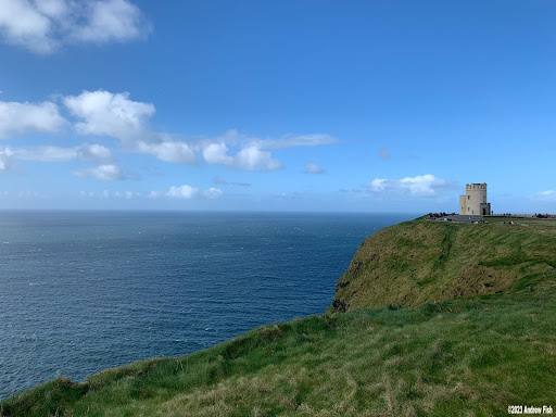 A stone tower stands on a grassy cliff overlooking the blue ocean under a bright sky with scattered clouds.
