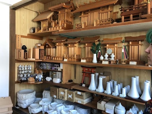 Wooden shelves display miniature Shinto shrines, ritual objects, white ceramic vessels, small torii gates, and various religious ornaments. A person with green hair is partially visible on the right.