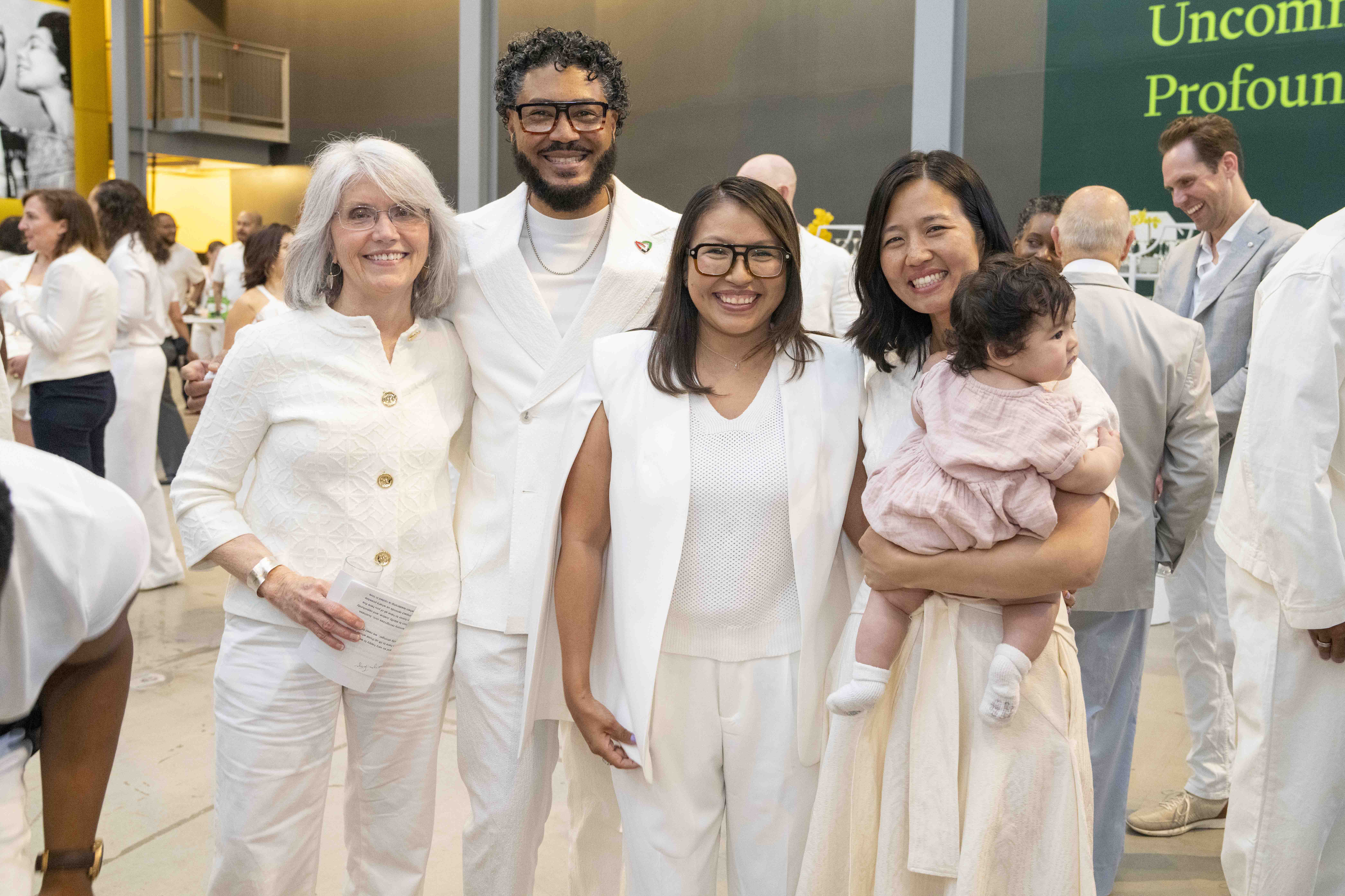Four adults in white outfits stand together, smiling at an indoor event. One woman, holding a baby, radiates warmth. Other attendees in white mingle nearby, their joyful resistance to routine sparking imagination amid green signage on the wall.