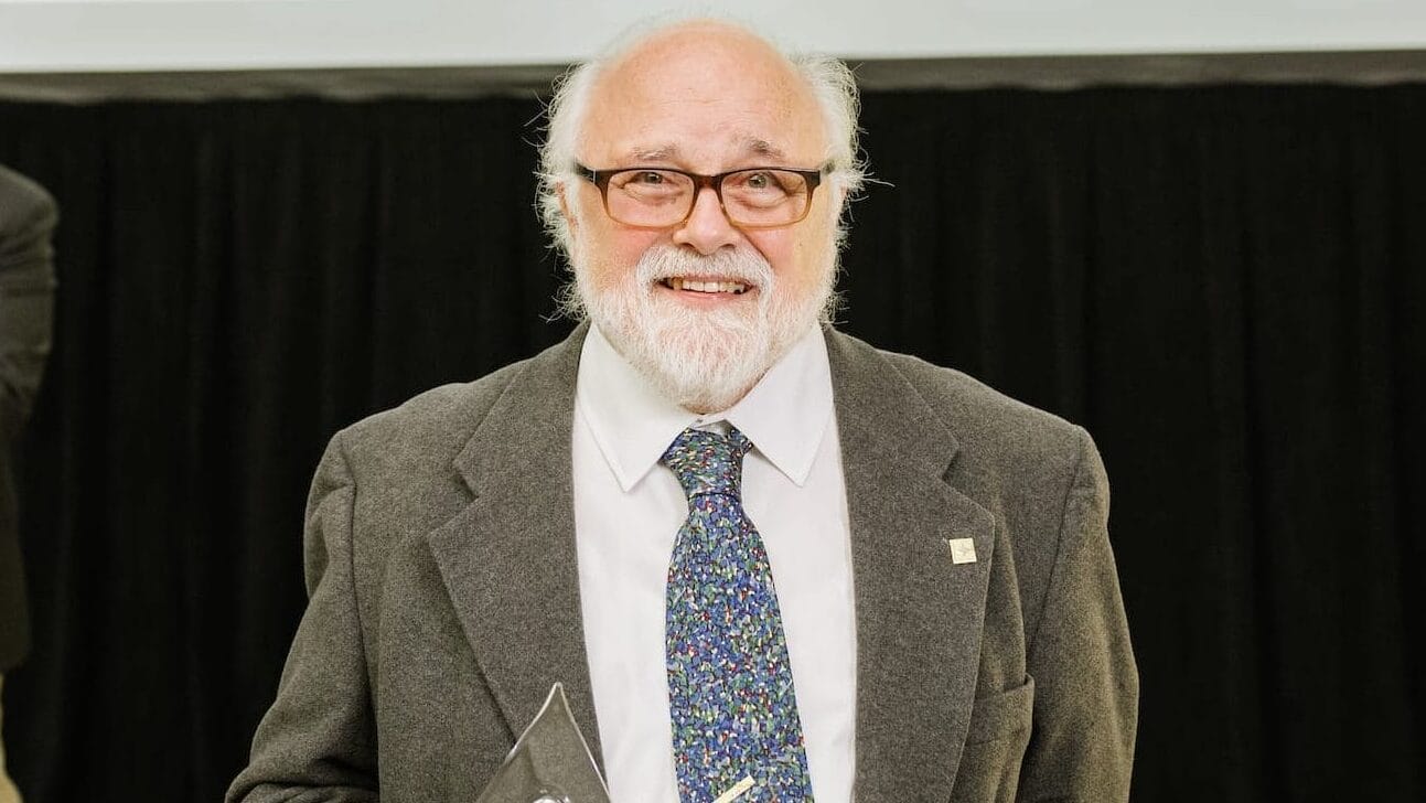 An older man with white hair and a beard, wearing glasses, a suit, and a patterned tie, smiles while holding his Common Good Award in front of a large 150 Years MassArt Common Good Awards sign.