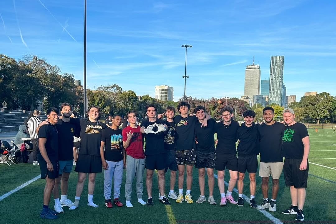 A group of thirteen young people stand together on a grass sports field, smiling at the camera. Dressed in athletic clothing, they gather for some recreation with a city skyline and trees behind them under a clear blue sky.
