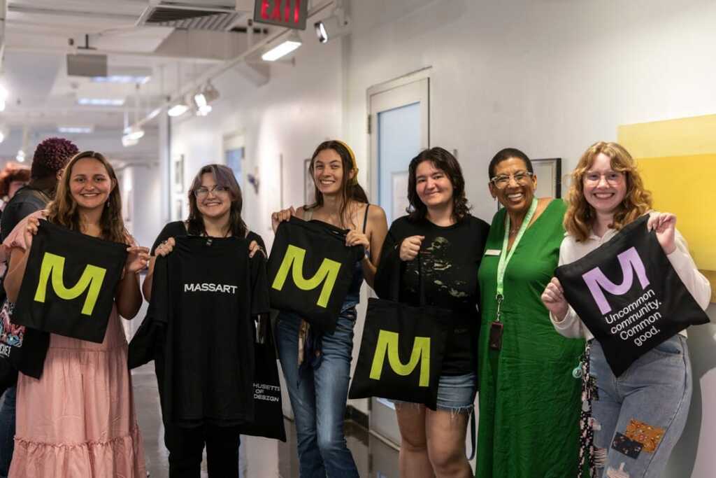 Six people stand in a brightly lit hallway, smiling and holding black tote bags with a large green or purple “M” logo. One bag reads 91 Supply, while another says Uncommon Community Cohort, suggesting creative books or materials inside.
