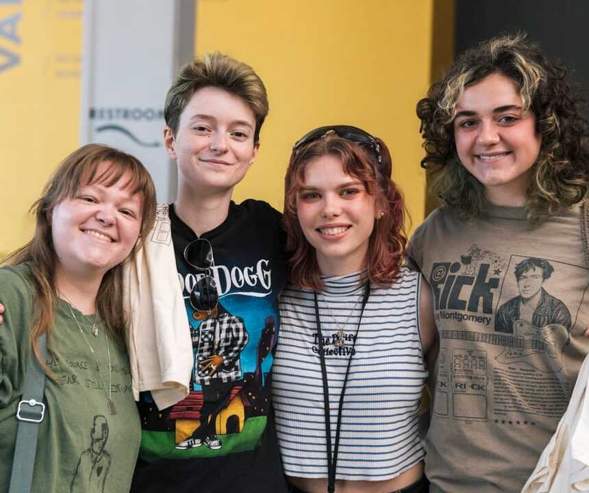 Four smiling young adults stand close together, posing for a photo. They wear casual clothing, including graphic t-shirts, and appear happy and relaxed in an indoor setting with a yellow and gray background.