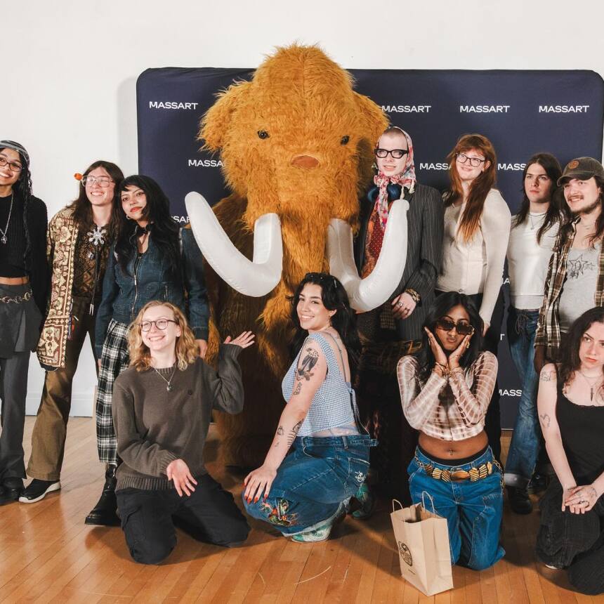 A group of people pose and smile around a large, furry mammoth mascot with big tusks in front of a MassArt backdrop. The group is indoors on a wooden floor.