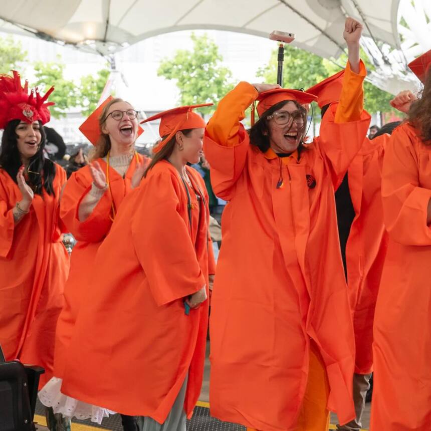 A group of graduates in orange caps and gowns cheer and celebrate while walking in a graduation ceremony. One graduate wears a red feathered headdress. The event takes place under a large tent.