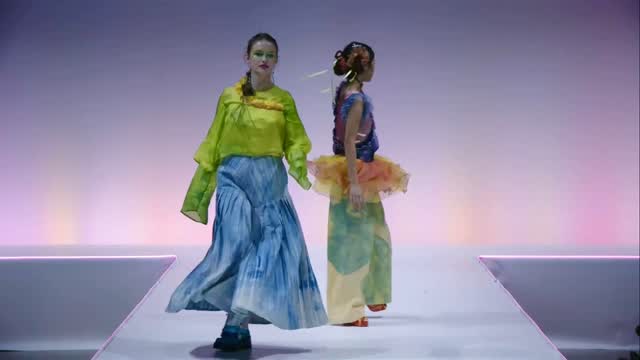 Two models walk on a runway wearing colorful, avant-garde outfits. The model on the left wears a sheer neon green top and blue skirt, while the one on the right wears a blue top with an orange tutu over pastel pants.