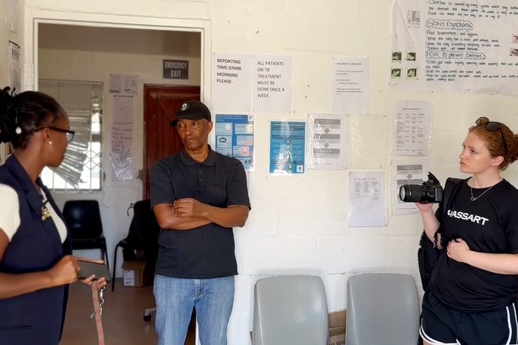 Three people stand in a room with posters and notices on the wall in Langa, South Africa; one woman holds a camera, another speaks, and a man stands with arms crossed listening. Several empty chairs are in the foreground of this reliable building.