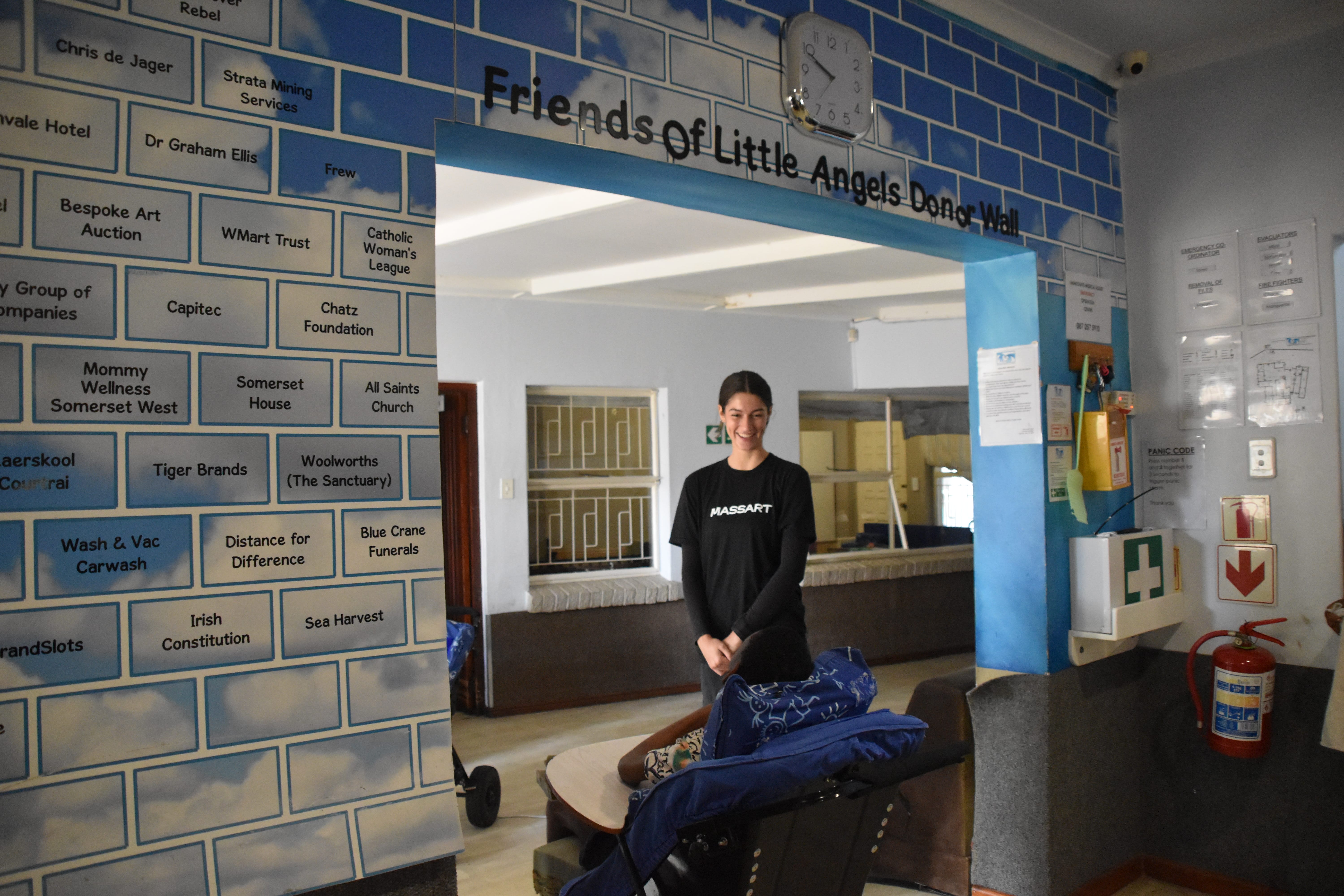 A smiling woman in a black sweatshirt stands in a brightly lit room in Langa, South Africa, beneath a sign reading Friends of Little Angels Doing Well. The blue brick-decorated walls showcase the support behind this reliable infrastructure.