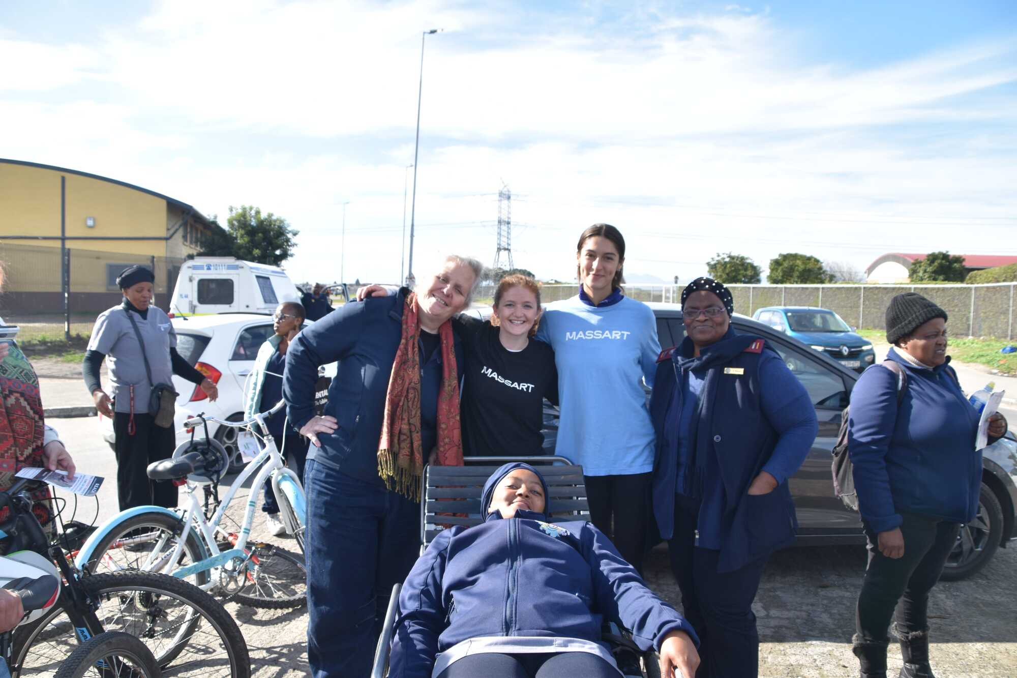 Four women stand smiling outdoors, with one lying on a bench in the foreground. Two of the standing women wear MASSART shirts, highlighting their pride as MassArt students. Other people, bicycles, and cars fill the sunny background.