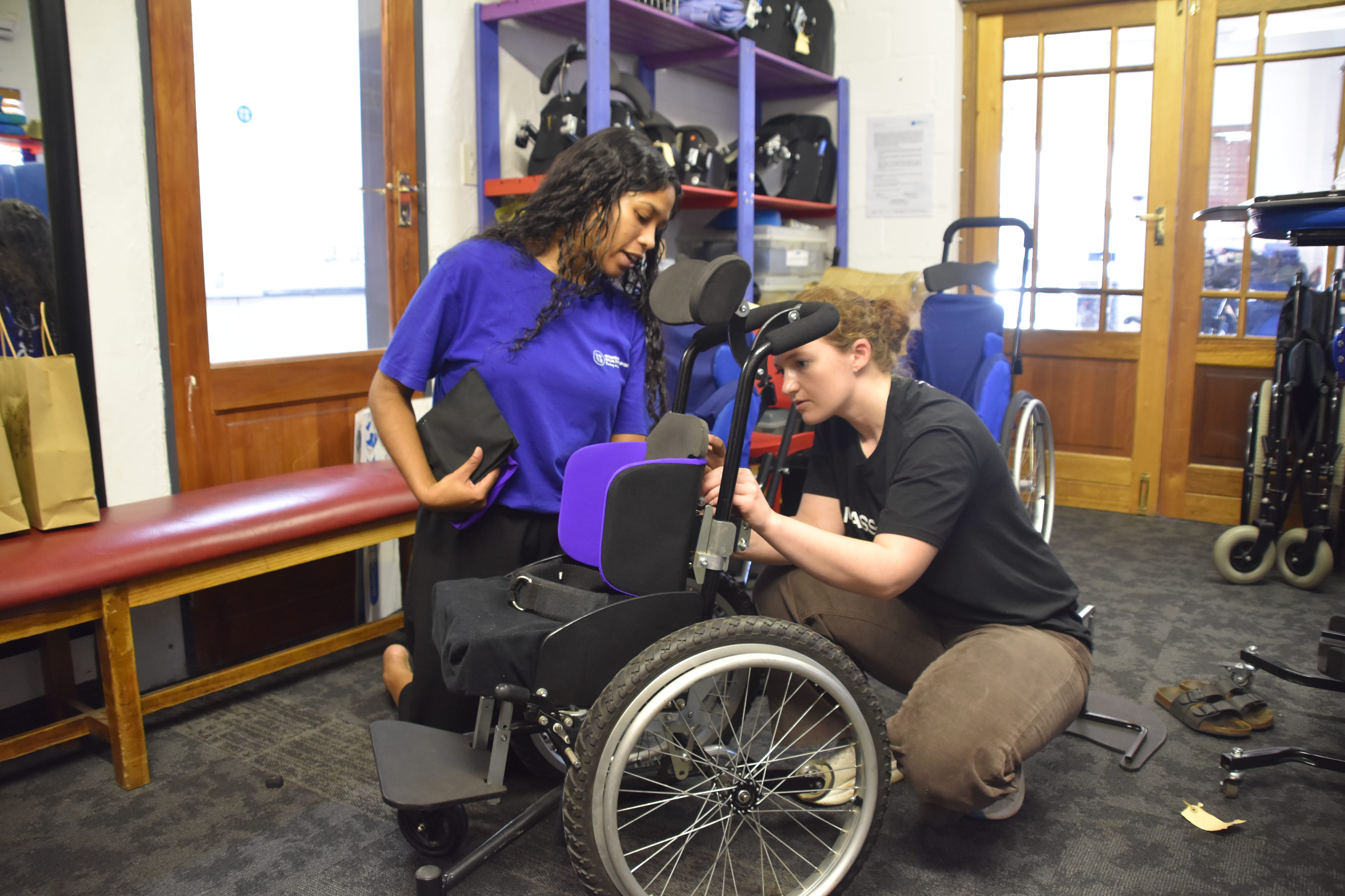 Two women in Langa, South Africa, adjust a specialized wheelchair together in a room with equipment and shelves. One kneels to work on the chair as the other observes, both focused on ensuring reliable emergency access.
