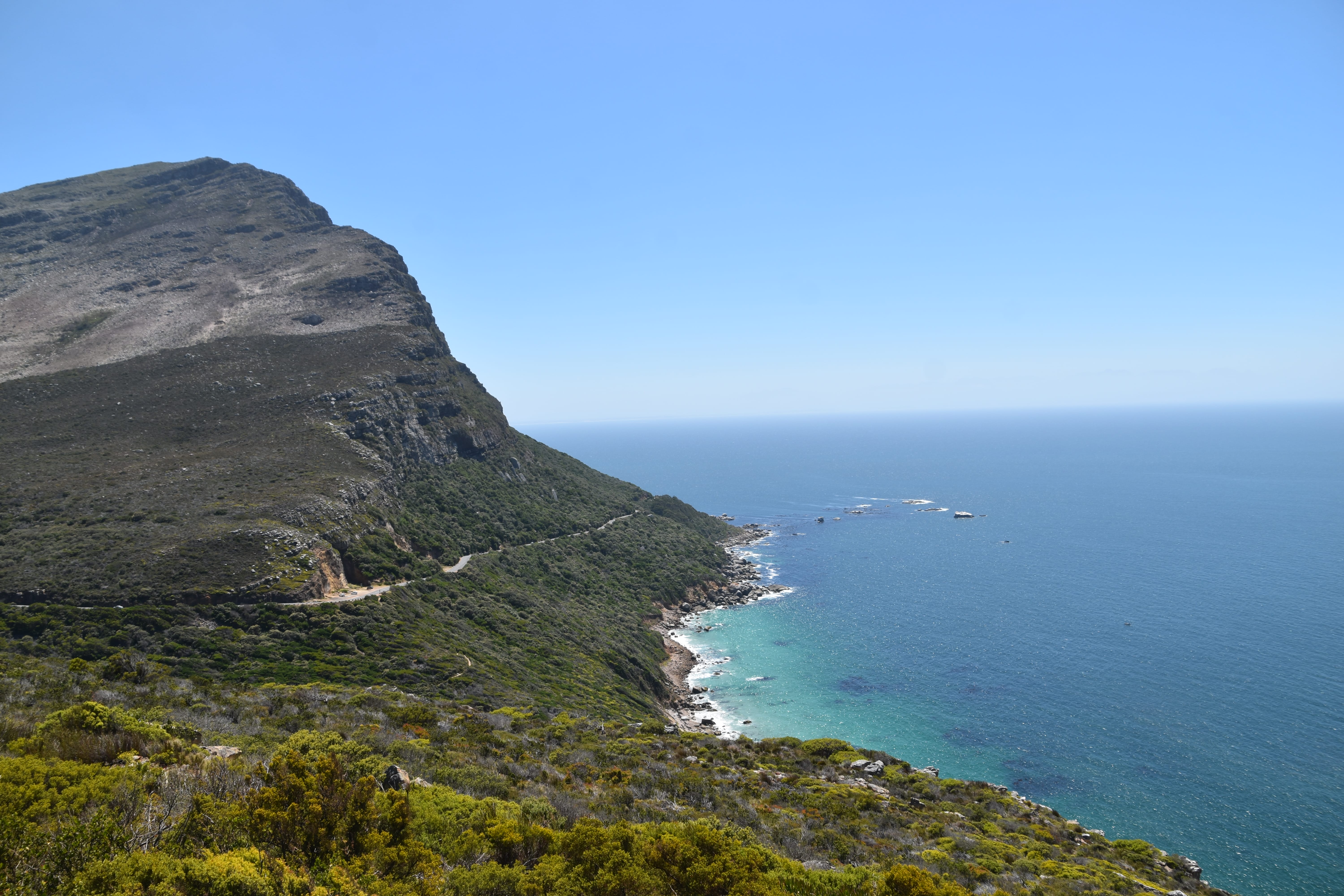 A rugged coastline in South Africa with a winding road hugs a steep, rocky hill. Green vegetation covers the foreground, while turquoise ocean waves meet the rocky shore under a clear blue sky, offering easy emergency access along the route.