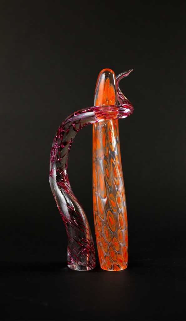 Two tall, abstract glass sculptures stand against a black background. One clear piece features orange and yellow oval patterns; the other, with red streaks, curves around the first—evoking the feeling of Growing Together through glass.
