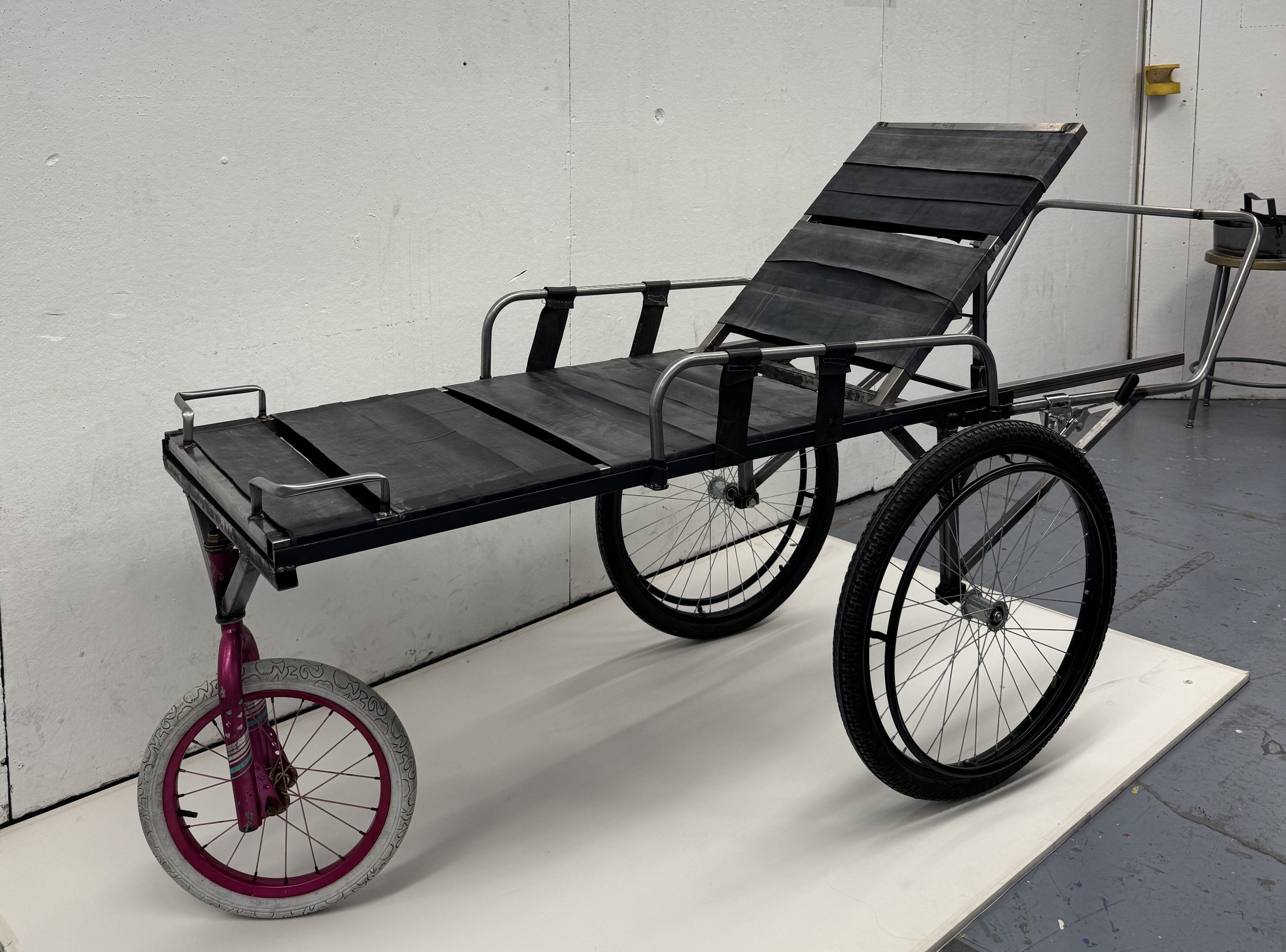 A three-wheeled, black metal lounge chair with a reclining backrest and armrests, featuring two large rear wheels and a small front wheel with a pink rim, designed for emergency access in environments like Langa South Africa.
