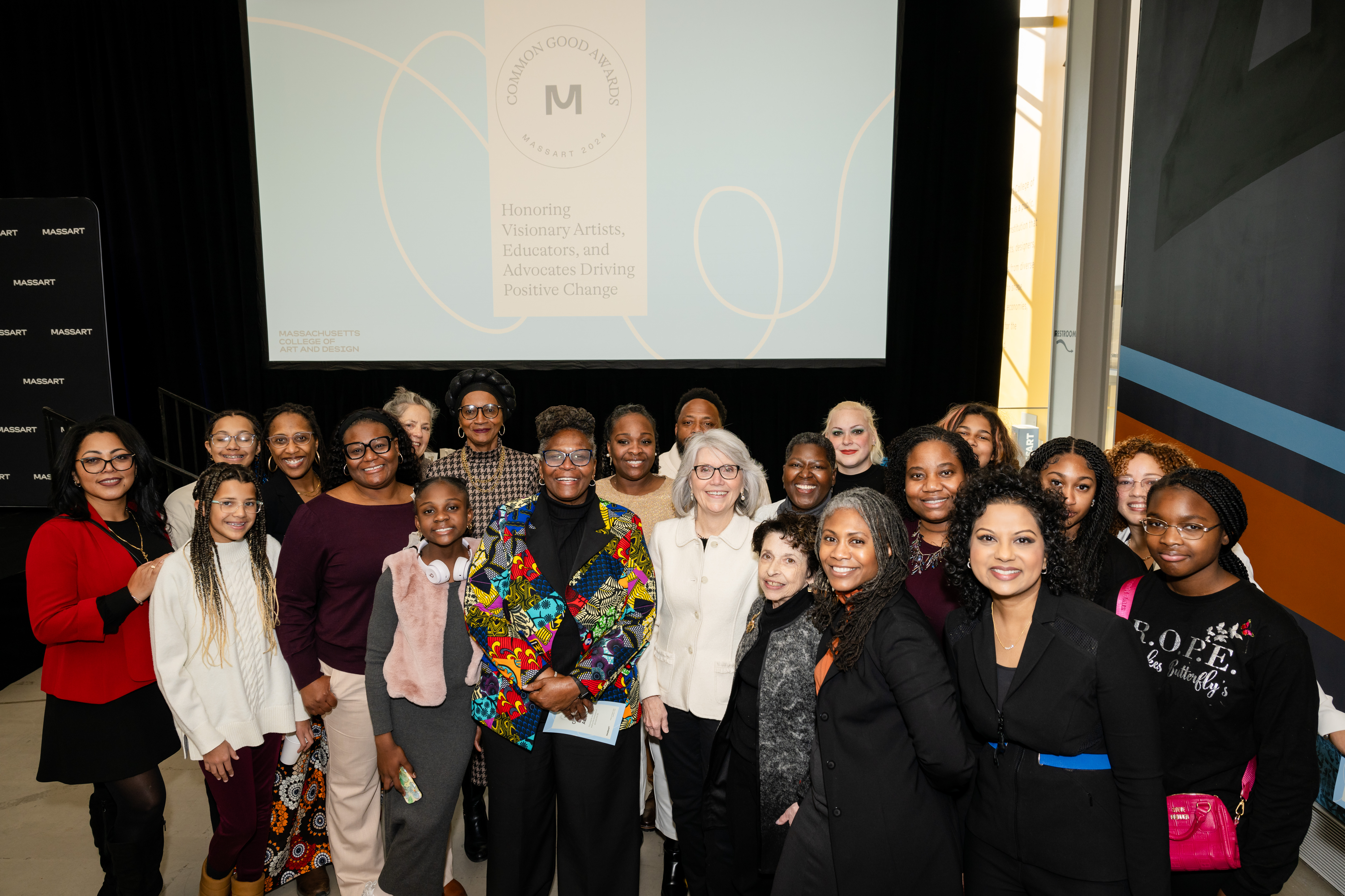 A diverse group of women and girls pose together, smiling for a group photo at an indoor event. A large screen in the background displays a message honoring 2025 MassArt Common Good Awards nominations for artists, educators, and advocates driving positive change.