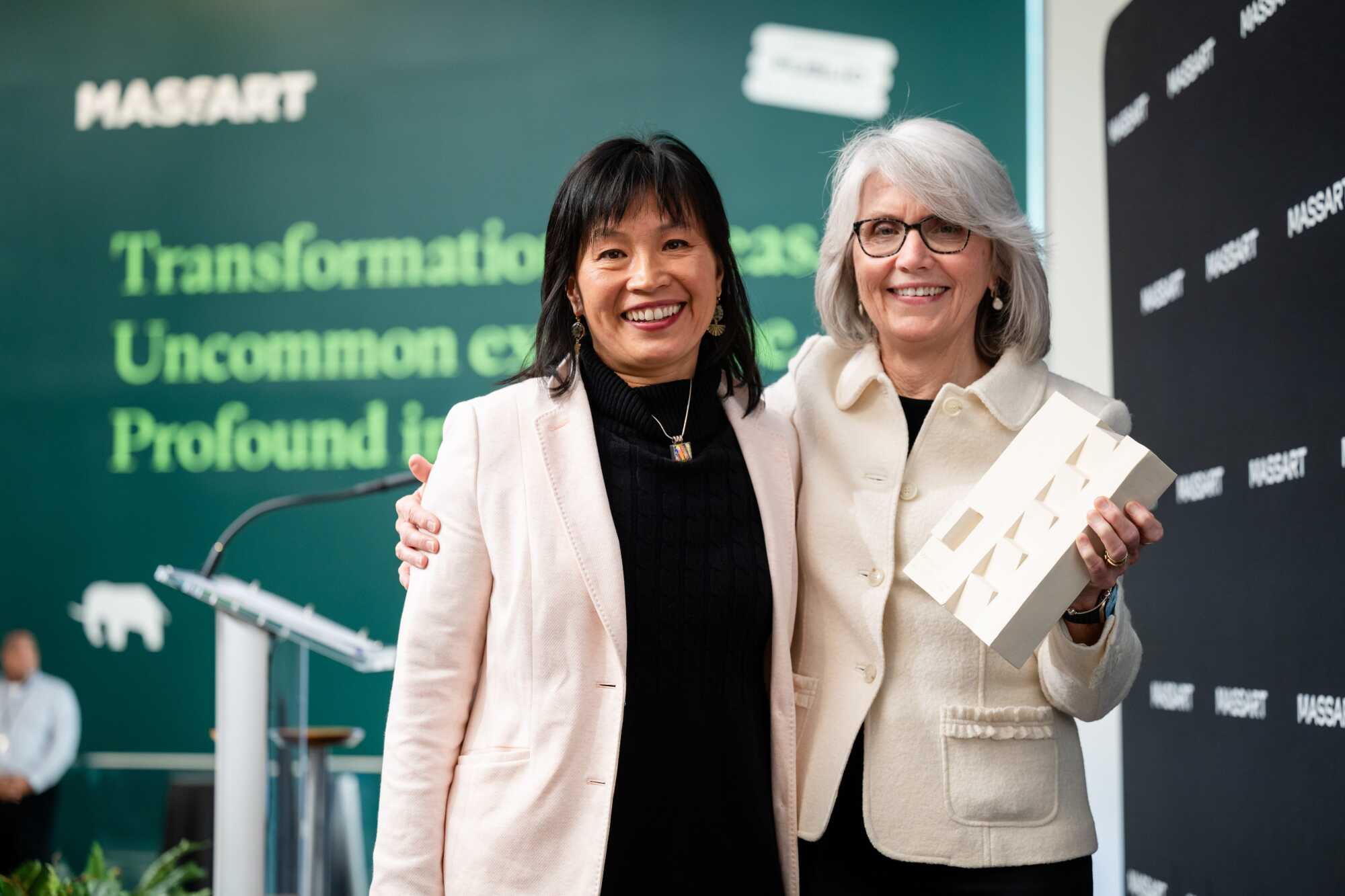 Two women smiling and standing together, one holding a white abstract Common Good Award. They are in front of a green wall with MASsART branding and text about transformation and profound impact, celebrating award recipients for the common good.