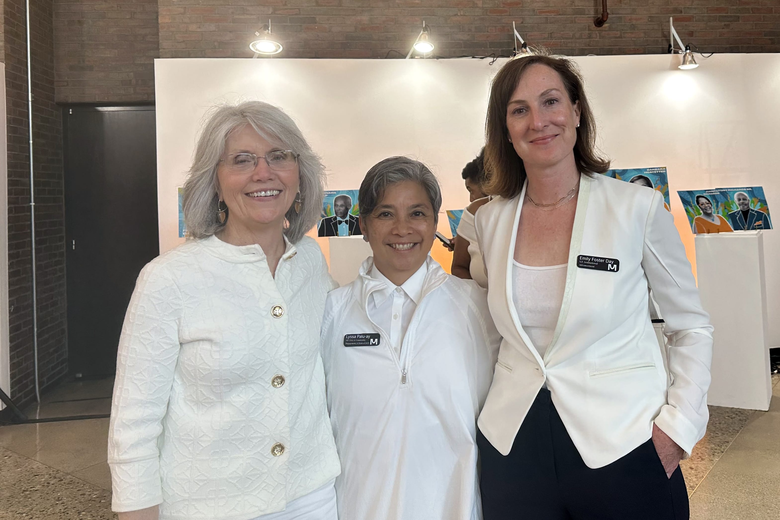 Three women wearing white tops and name badges stand smiling together indoors, with colorful art—showcasing imagination—displayed on the white wall behind them and a brick wall partially visible.
