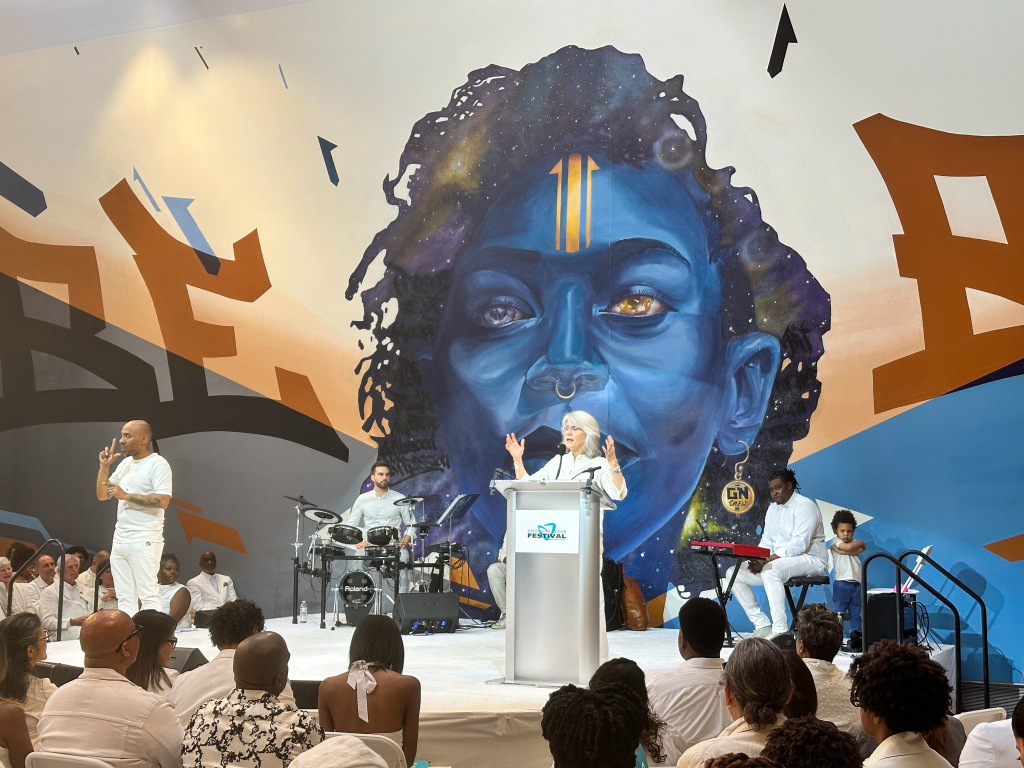 A woman speaks at a podium on stage, backed by musicians, in front of a vibrant mural of a blue-faced person. The audience, all dressed in white, listens attentively amid an atmosphere pulsing with imagination and artistry.