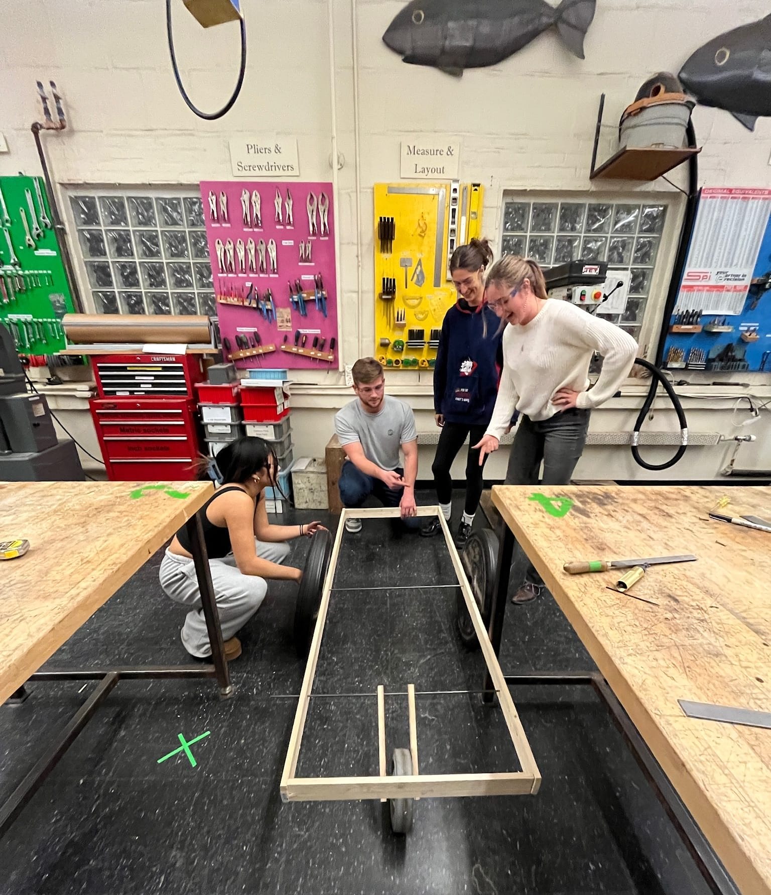 In a workshop in Langa, South Africa, four people collaborate around a rectangular wooden frame with wheels, surrounded by tools, diagrams, and bright tool boards. Their project focuses on improving Emergency Access in the community.